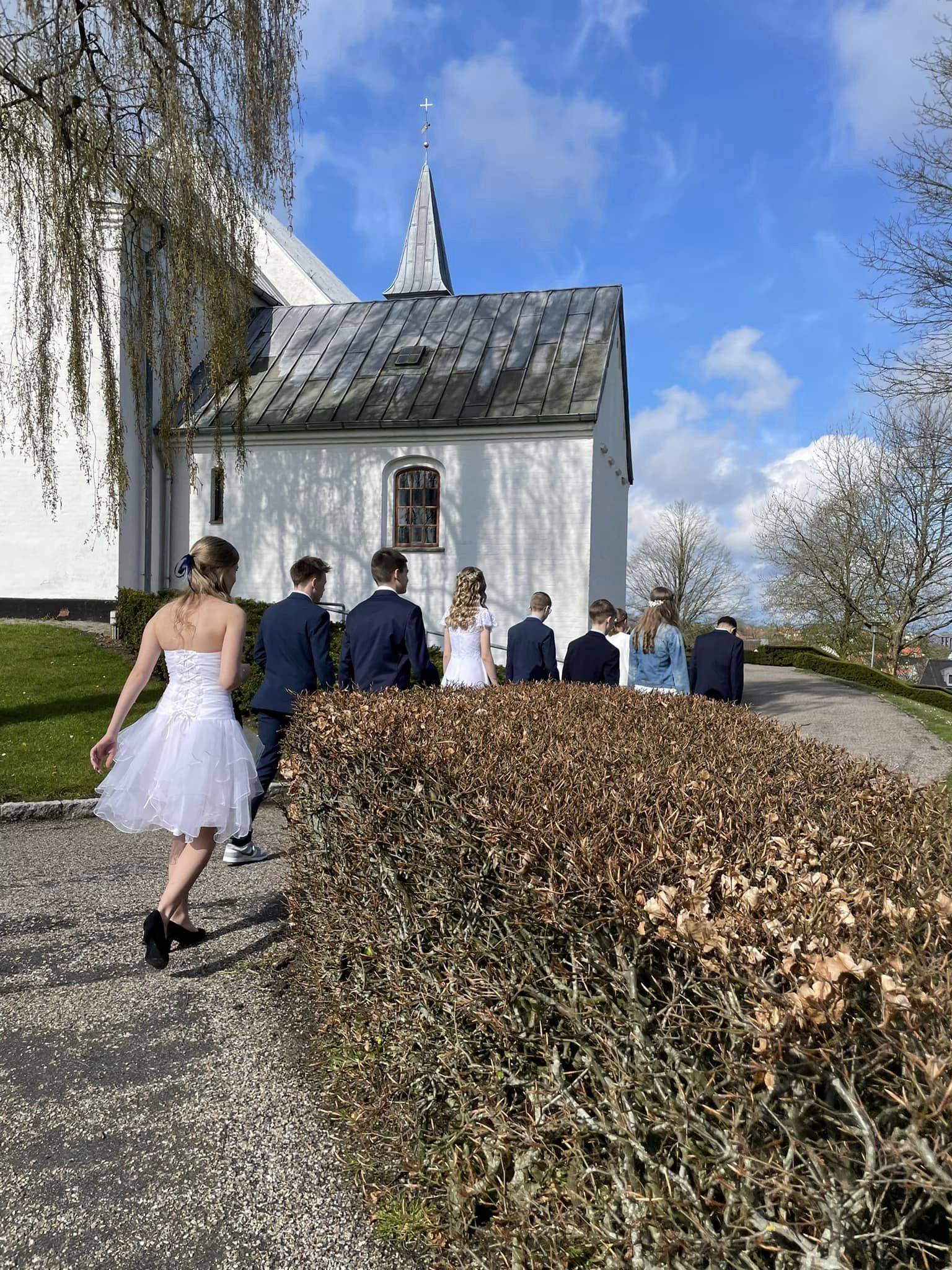 Ni unge mennesker i konfirmationstøj set fra ryggen på vej op mod kirke