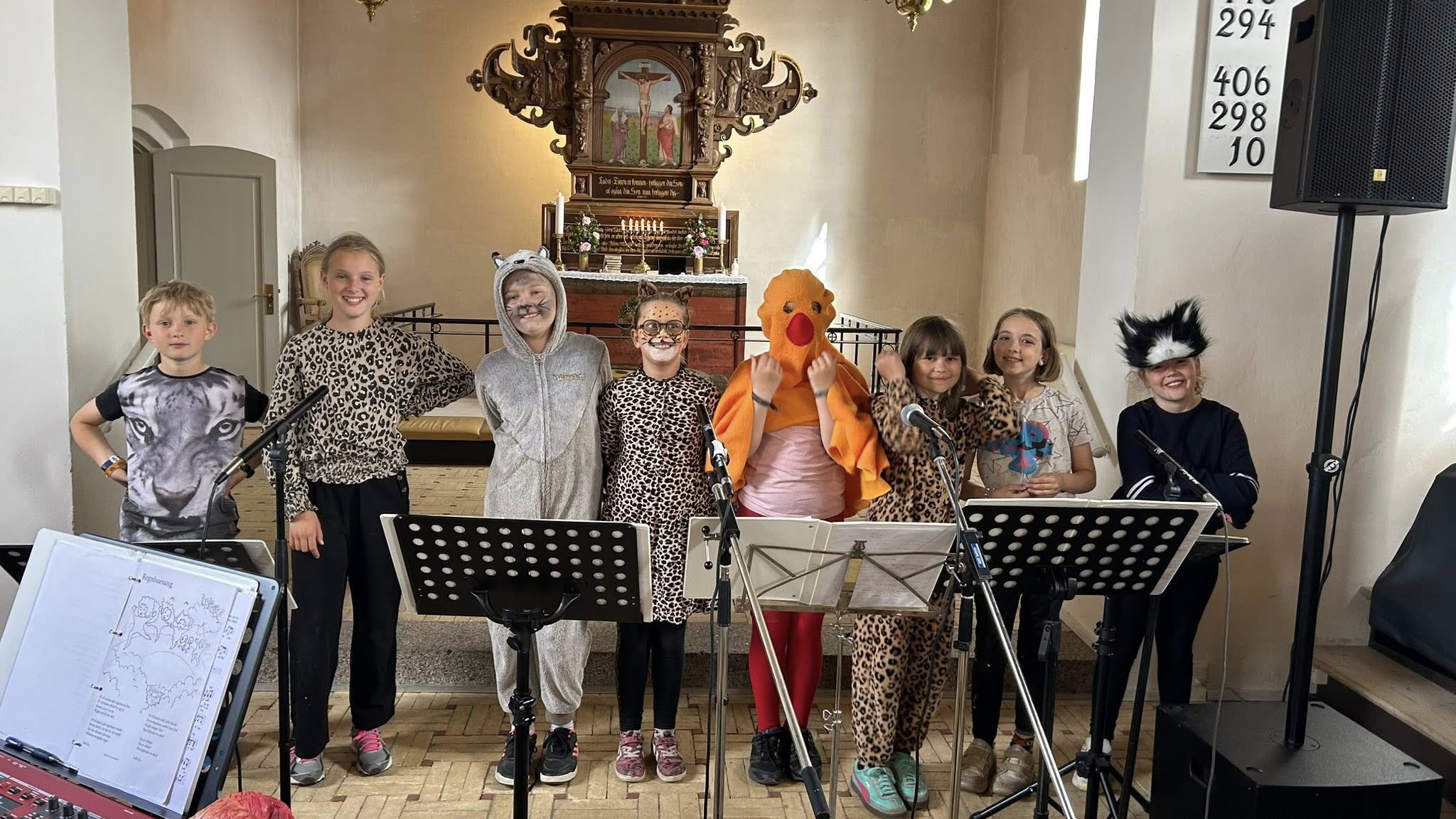 Otte udklædte børn stående i kirkekor med mikrononer og nodestativer