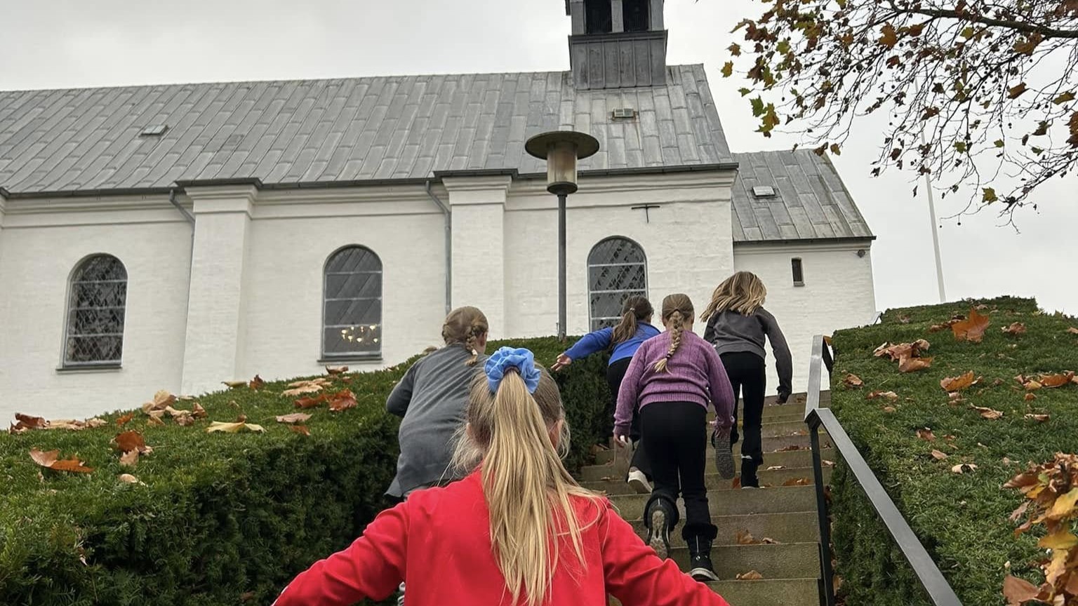 Børn set fra ryggen løbende op ad trappen mod en hvid kirke