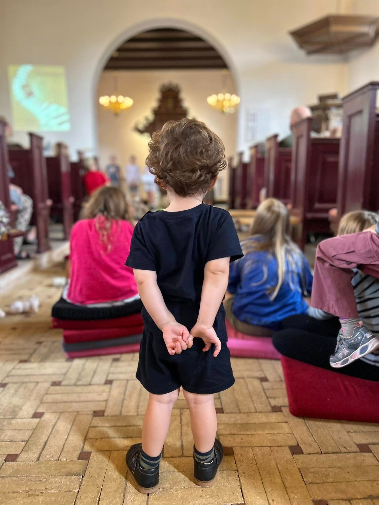 Børn set fra ryggen siddende i midtergang i kirkerum. Stående lille dreng i forgrunden. 