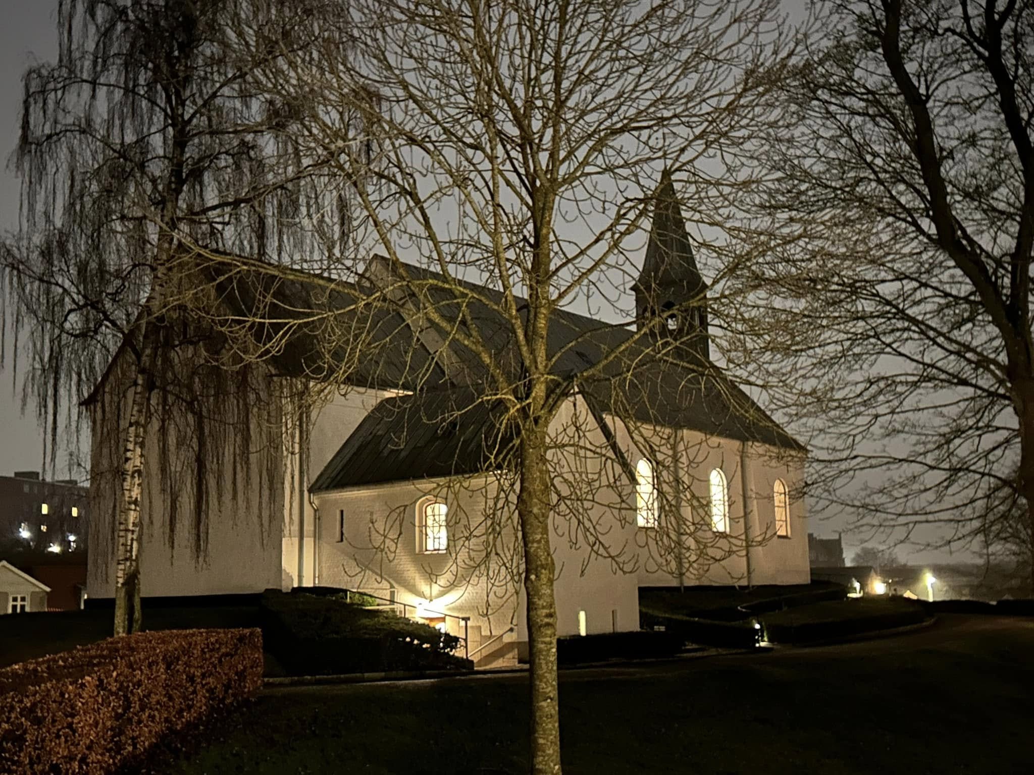 Oplyst hvid kirke i tusmørke omgivet af efterårstræer